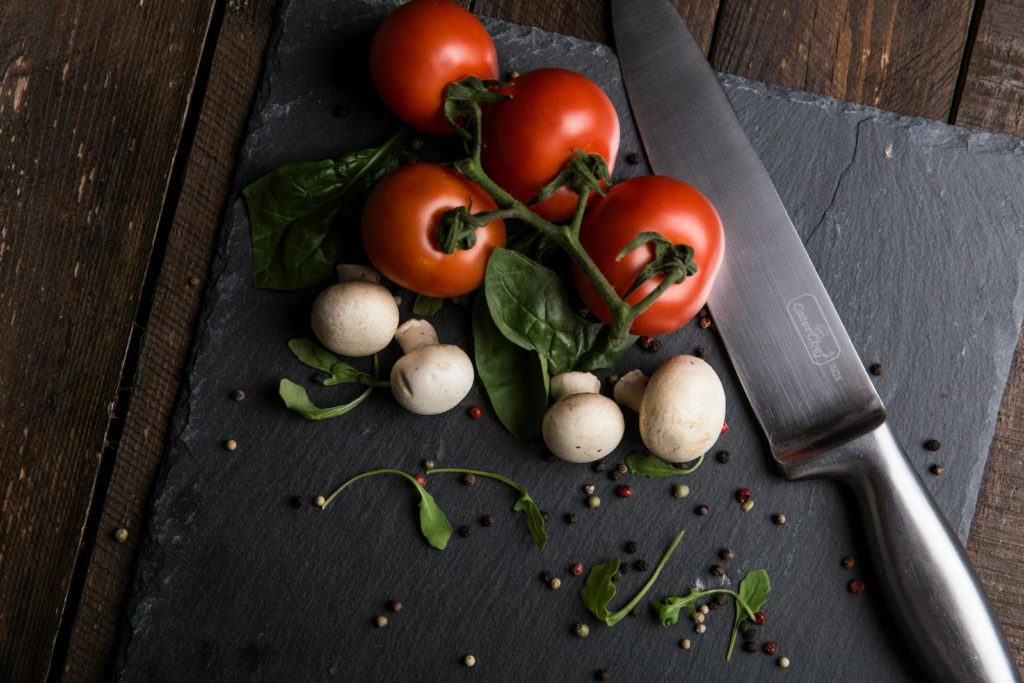 Top view of fresh tomatoes, mushrooms, and greens on a slate board with knife.